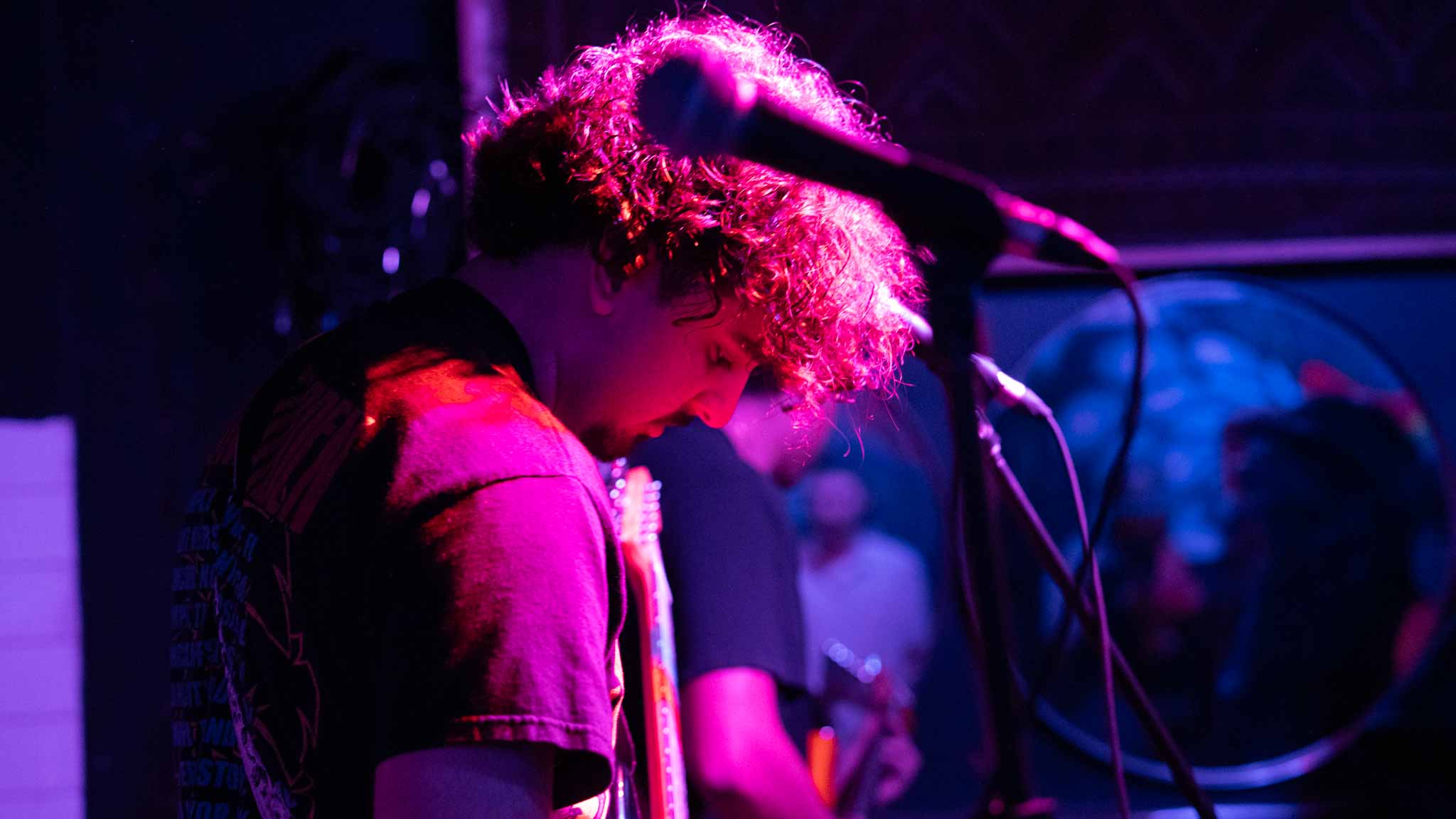 Musician bowed over a guitar beneath a microphone and red stage light
